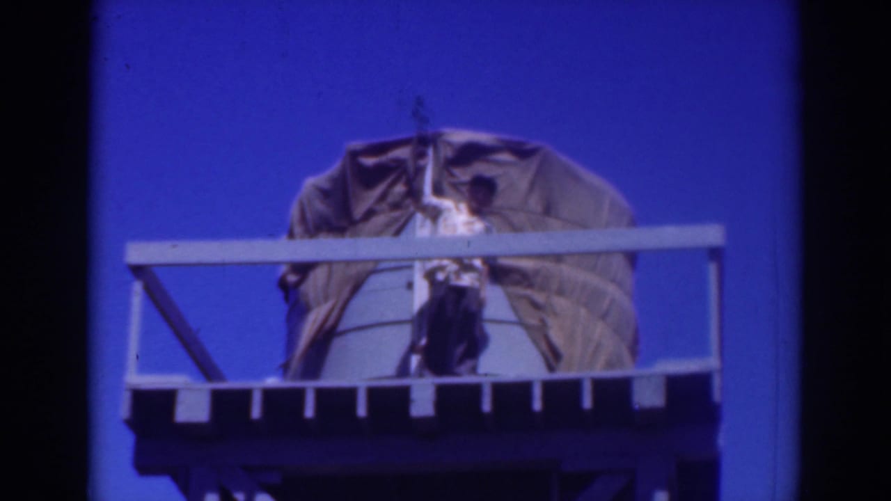 1937 Sacramento 8mm frame showing a man on a railed water tank platform adjusting a large tarp against a vivid blue sky.