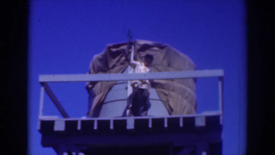 1937 Sacramento 8mm frame showing a man on a railed water tank platform adjusting a large tarp against a vivid blue sky.