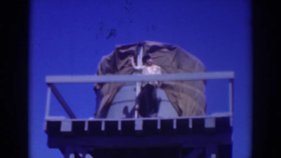 A distant 1937 Sacramento home-movie view shows a man on a platform, partly behind railings, working with a tarp over a water tank.