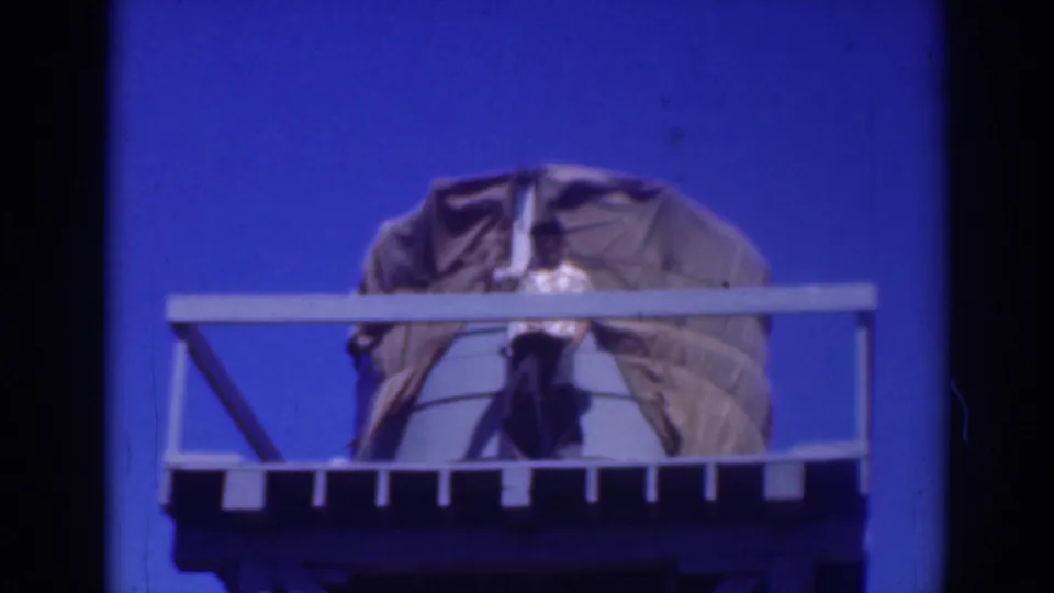 1937 Sacramento 8mm frame of a man on a high railed platform with a tarp-draped water tank behind him.