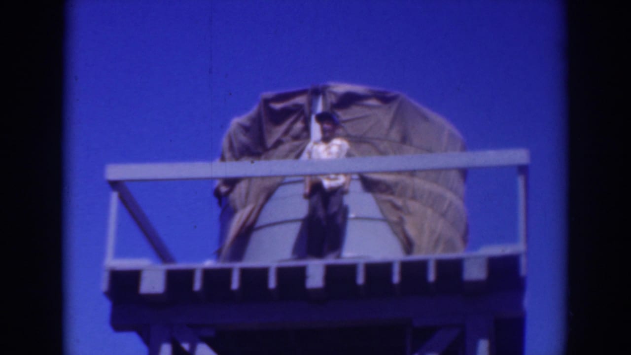 Man in a hat stands on an elevated platform beside a tarp-covered tank against blue sky.