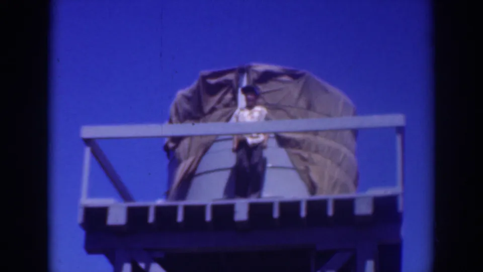 In 1937 Sacramento footage, a man stands on a raised water-tank platform near a large tarp-covered tank under a vivid blue sky.