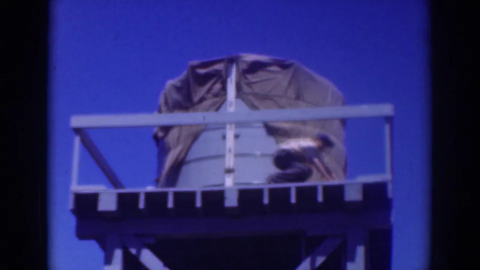 The man crouches on the elevated platform while working near the covered tank, seen in soft 8mm color with visible frame borders.