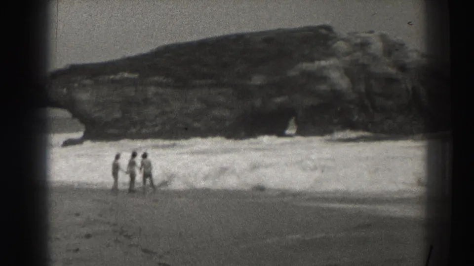 Three distant people walk along the 1937 Santa Cruz shoreline with surf and a large coastal rock formation behind them.