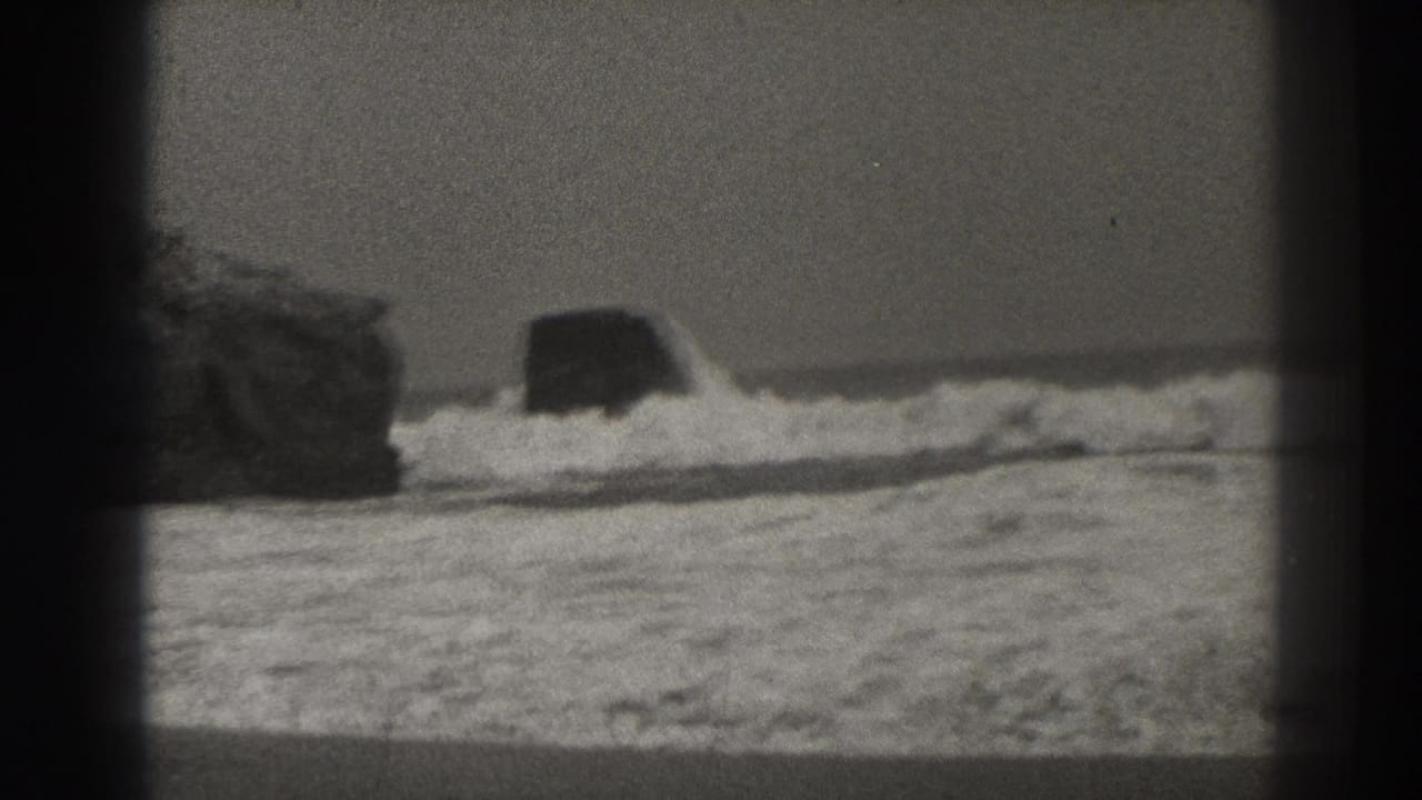 Waves break around dark rocks near a flat ocean horizon.