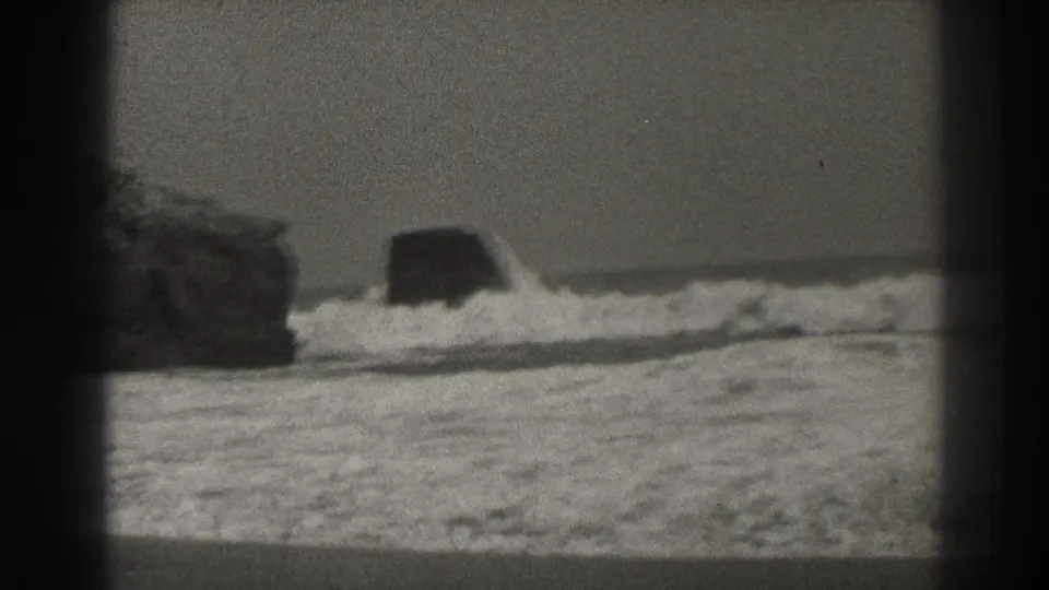 1937 Santa Cruz 8mm frame showing surf breaking against dark coastal boulders under a muted sky.