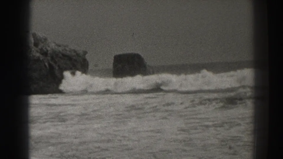 Santa Cruz coastal home-movie frame with a wave cresting beside dark boulders and a steady horizon.
