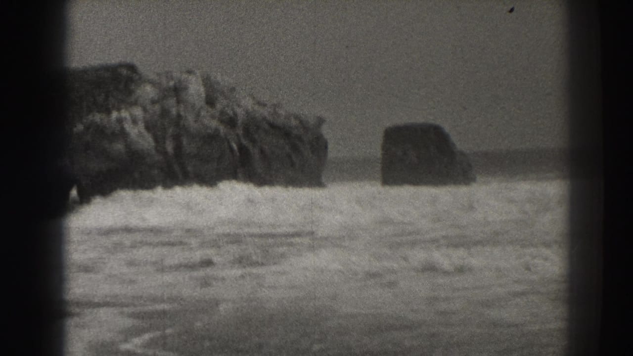 Black-and-white surf with large rocks offshore and waves breaking near the beach.