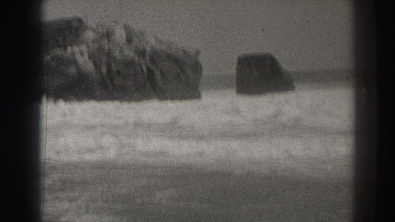 Rocky surf line with waves rolling toward shore in black-and-white footage.