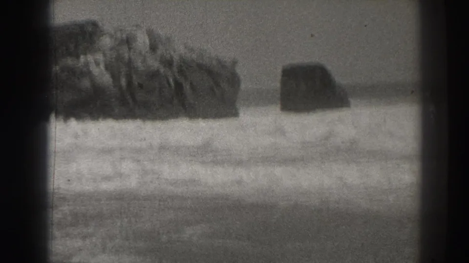 Black-and-white 8mm view of Santa Cruz surf, with large rocks and foamy waves moving across the shoreline.