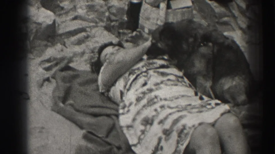 Santa Cruz beach home movie frame with a woman in patterned clothing sunbathing next to a resting dog on the sand.
