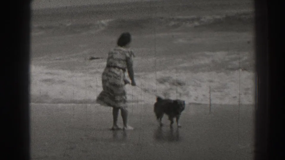 In 1937 Santa Cruz home-movie footage, a woman in a patterned dress stands with her dog on wet beach sand as waves break behind them.