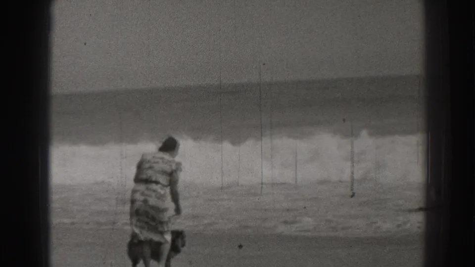 In 1937 Santa Cruz beach footage, a woman in a patterned dress walks a small dog along wet sand near breaking waves.