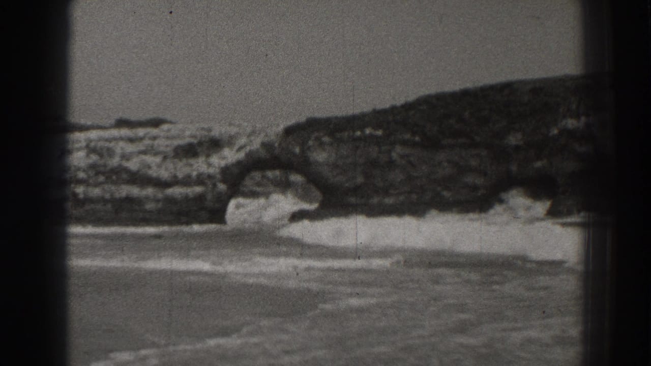White surf breaks against dark coastal rocks and a natural arch beside a sandy beach.