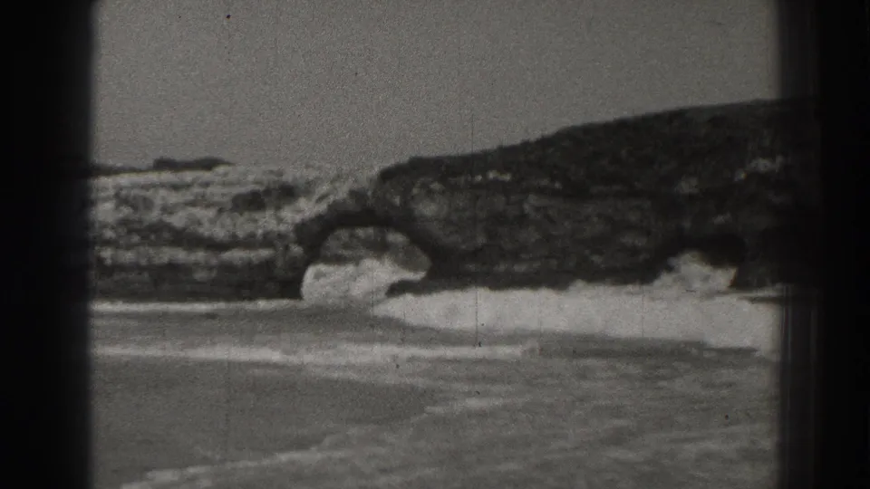 1937 Santa Cruz 8mm frame showing waves washing through a rocky sea arch near a sandy shoreline.