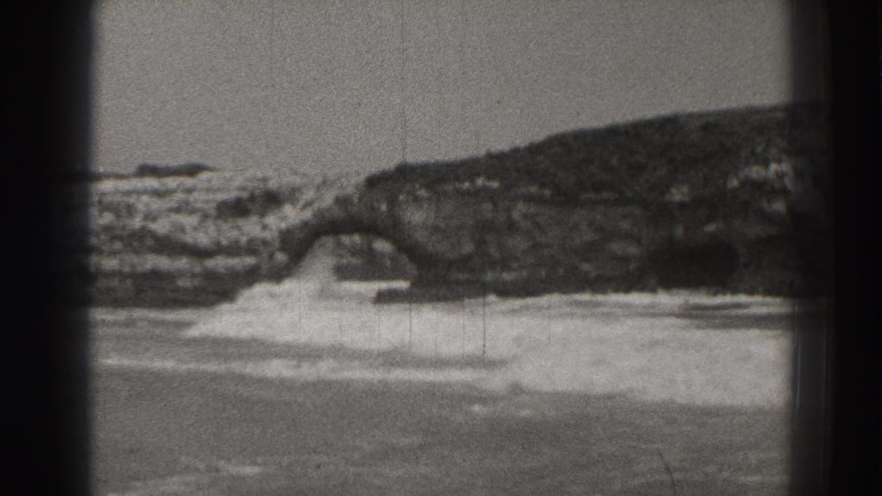 A wave crashes beneath a rock arch while foam spreads along the beach.
