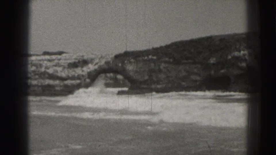 Black-and-white Santa Cruz coastal frame with foamy surf breaking under a dark rock arch in 1937 home-movie footage.