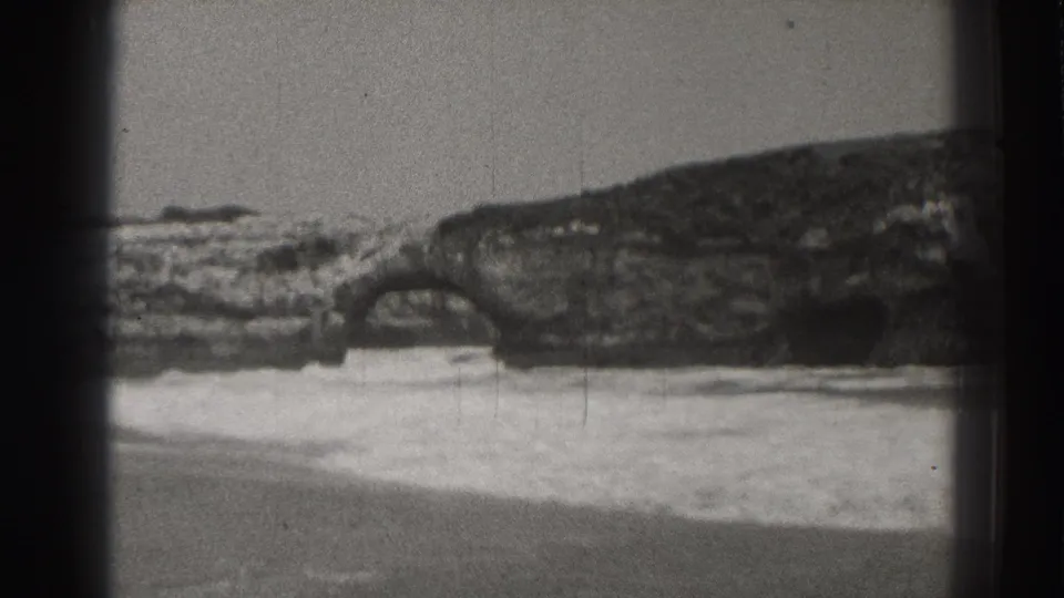 Santa Cruz shoreline frame from 1937 showing surf sliding over sand beneath a rugged coastal rock formation.