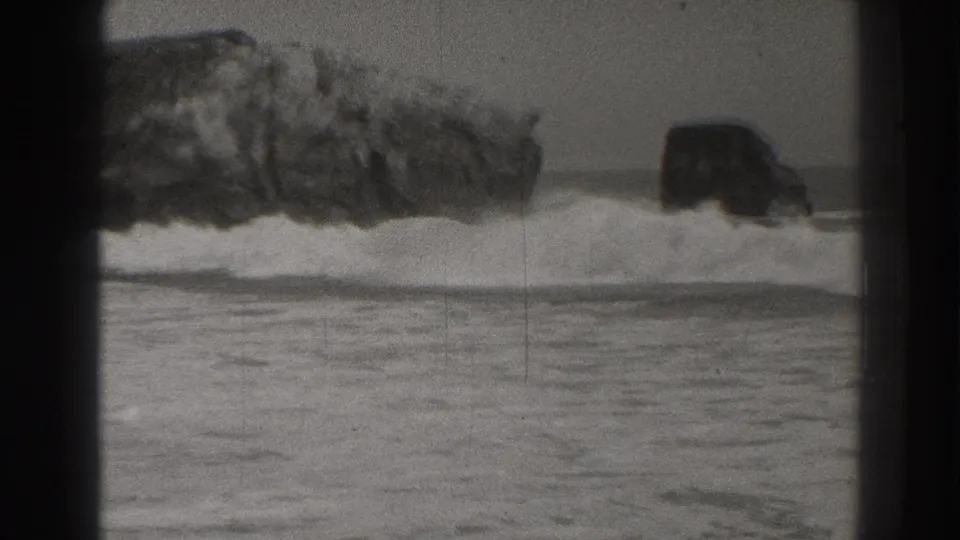Late-1930s Santa Cruz 8mm frame with surf breaking around a rocky outcrop and smaller offshore rock.