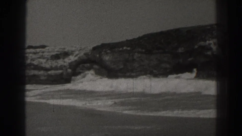 1937 Santa Cruz 8mm view of a rocky coastal formation with surf washing around its base and wet shoreline below.