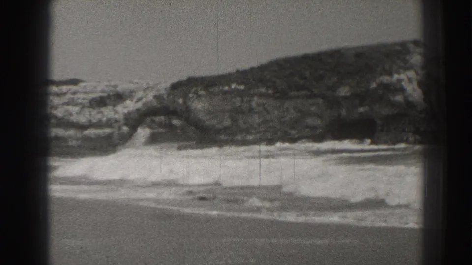 1937 Santa Cruz coastal frame showing a low rock formation with bright surf breaking around it and water stretching behind.