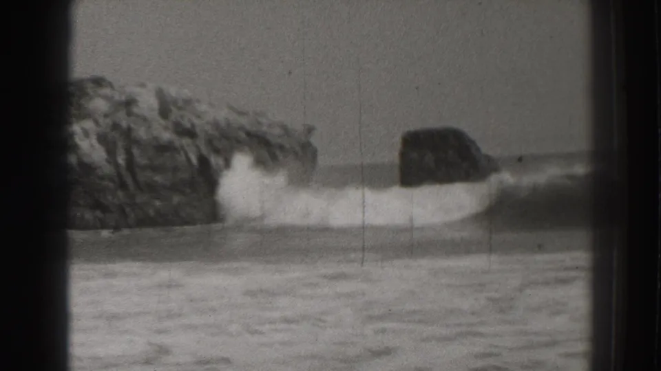 Black-and-white 8mm frame from Santa Cruz showing white surf moving around jagged rocks and a darker offshore rock shape.