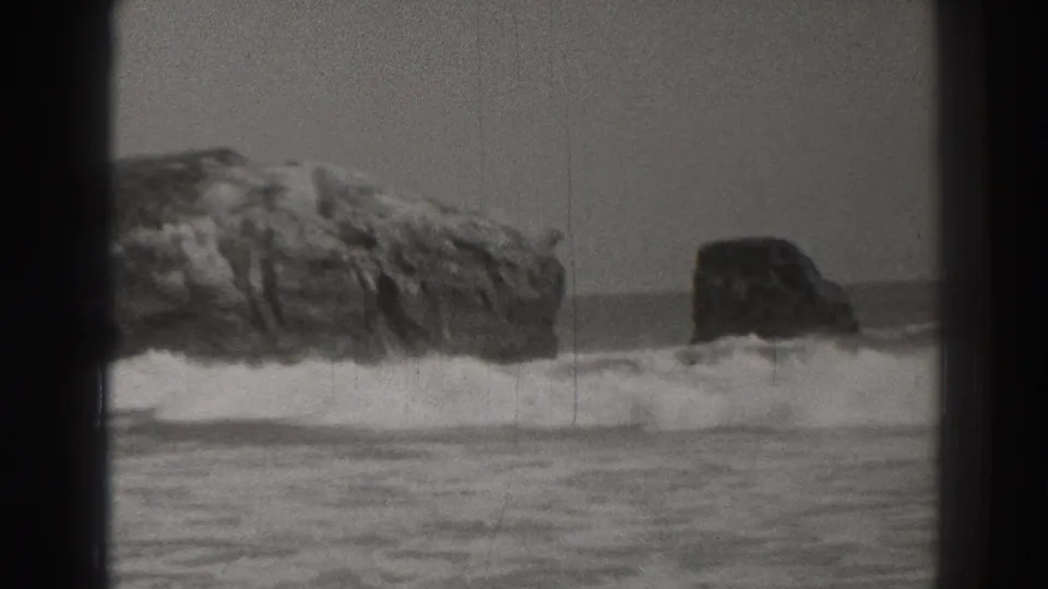 Grainy 8mm Santa Cruz coastal frame showing foamy surf between a jagged rock ledge and a dark rock offshore.