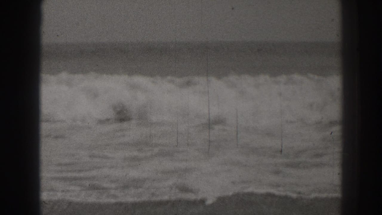 Foamy waves breaking near a flat shoreline under an overcast sky.
