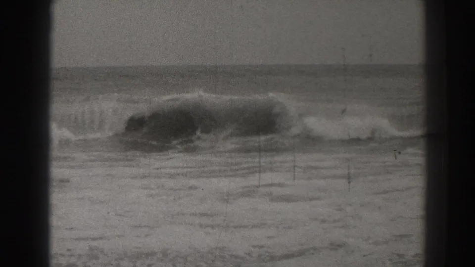 Black-and-white Santa Cruz surf footage shows a darker wave crest collapsing near shore, framed by visible 8mm borders and scratches.