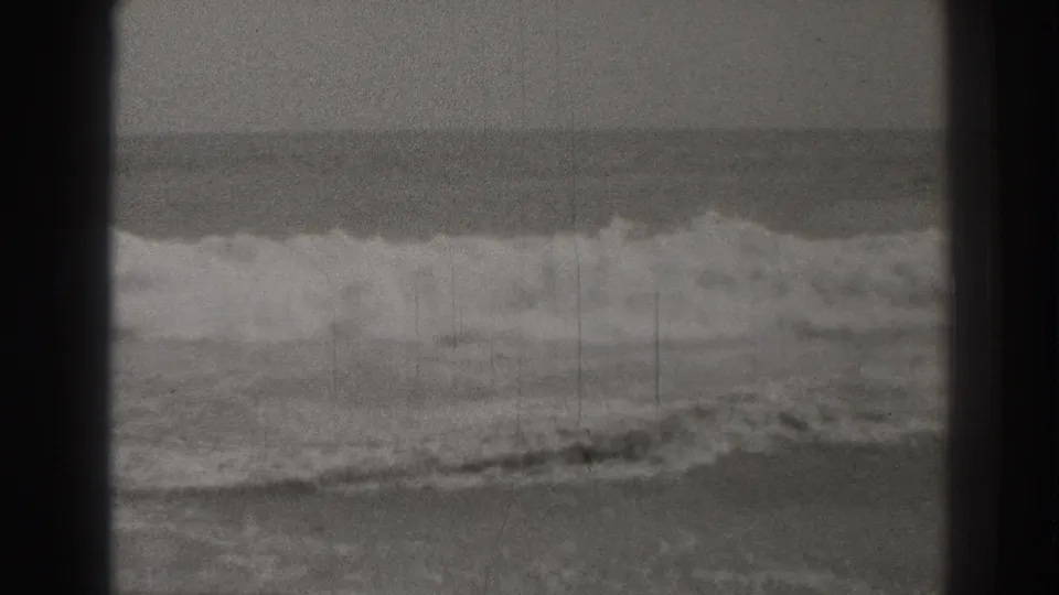 Black-and-white 8mm frame from Santa Cruz showing rolling ocean surf under a muted sky, with scratches and projector-edge shadowing.