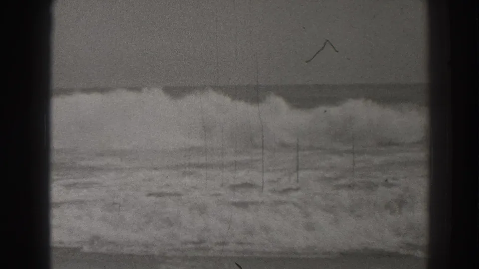Black-and-white Santa Cruz ocean view with surf breaking near shore, a level horizon, film grain, scratches, and a small mark on the frame.