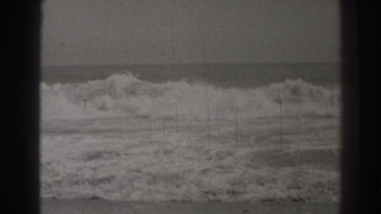 Breaking ocean waves under a gray sky, seen through dark 8mm frame edges.