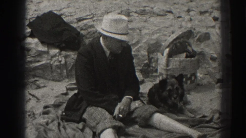 1937 Santa Cruz home-movie frame showing a seated man in a light fedora beside a dark dog on rocky ground.