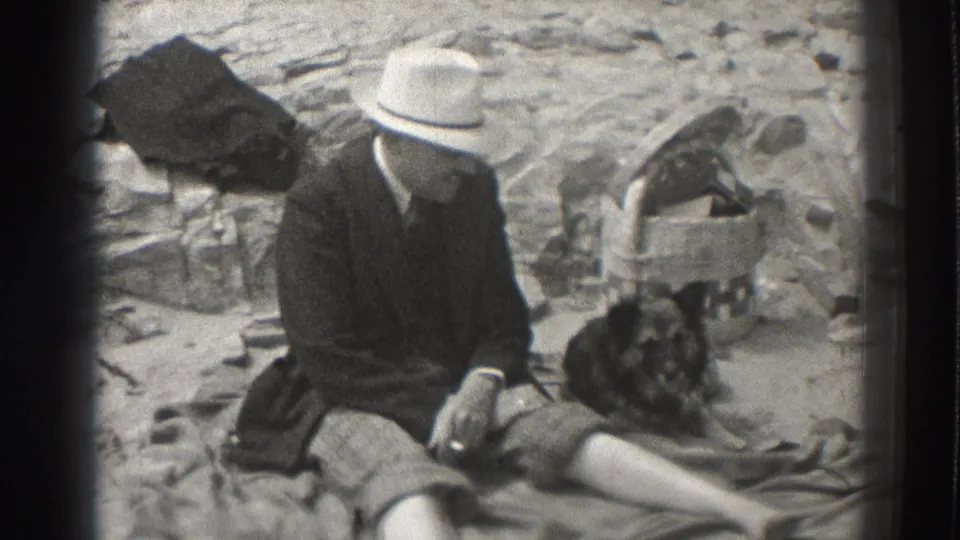 In Santa Cruz in 1937, a man in a light hat sits near a dark dog with rocks and a small container behind them.