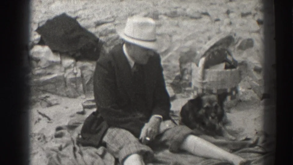 Black-and-white 8mm frame of a 1937 Santa Cruz outdoor pause, with a man in a fedora seated beside a dog.