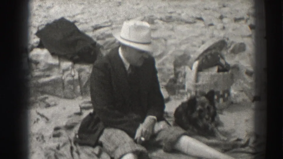 A 1937 Santa Cruz home-movie still shows a seated man in a light fedora beside a dark dog on sunlit rocks.