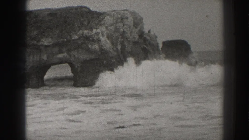 1937 Santa Cruz 8mm frame showing a natural rock arch, rugged sea stack, and white surf breaking below the cliff.