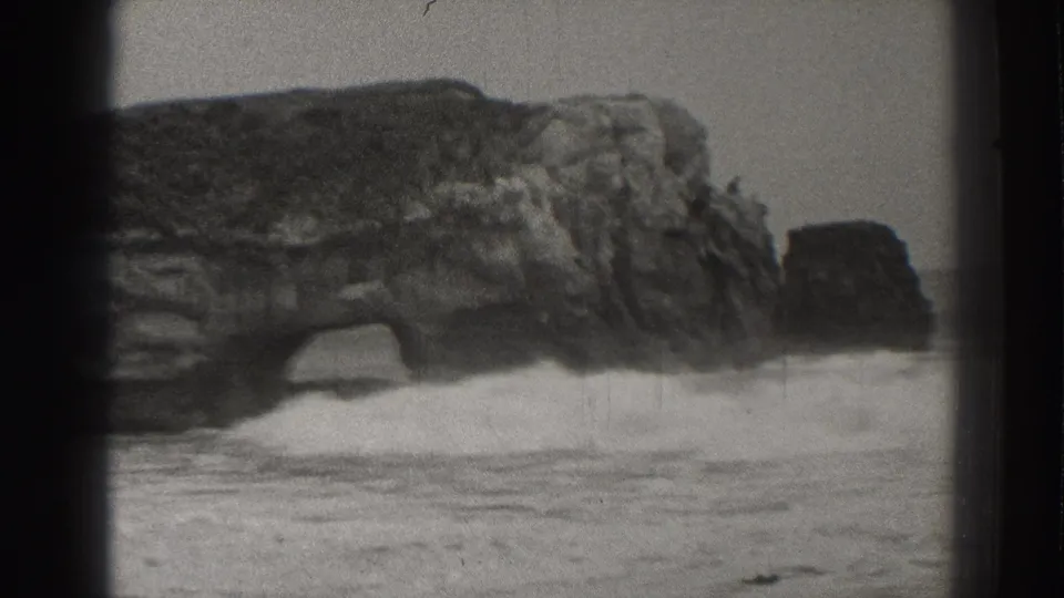 Black-and-white 8mm view of the Santa Cruz shoreline, with surf striking the rock arch and a separate sea stack in the distance.