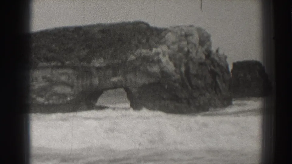 Final 8mm frame of the Santa Cruz rock arch, with the cliff face dominating the view and surf flattening into foam below.