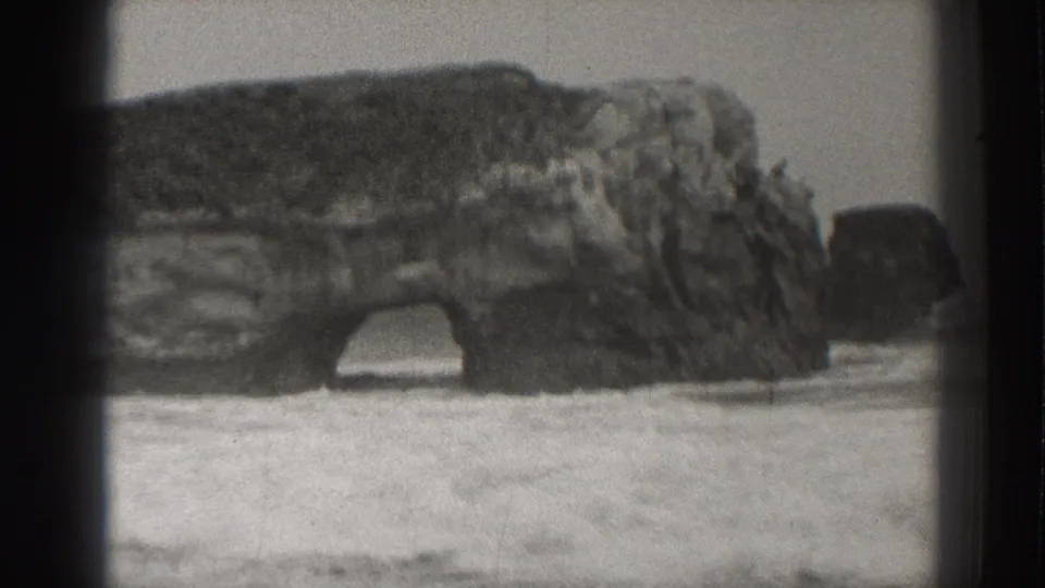 1937 Santa Cruz coastal view of a large rock arch as waves and sea foam move across the sandy shore.
