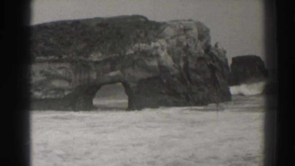 Black-and-white 8mm frame of the Santa Cruz sea arch with surf breaking and foam spreading toward shore.