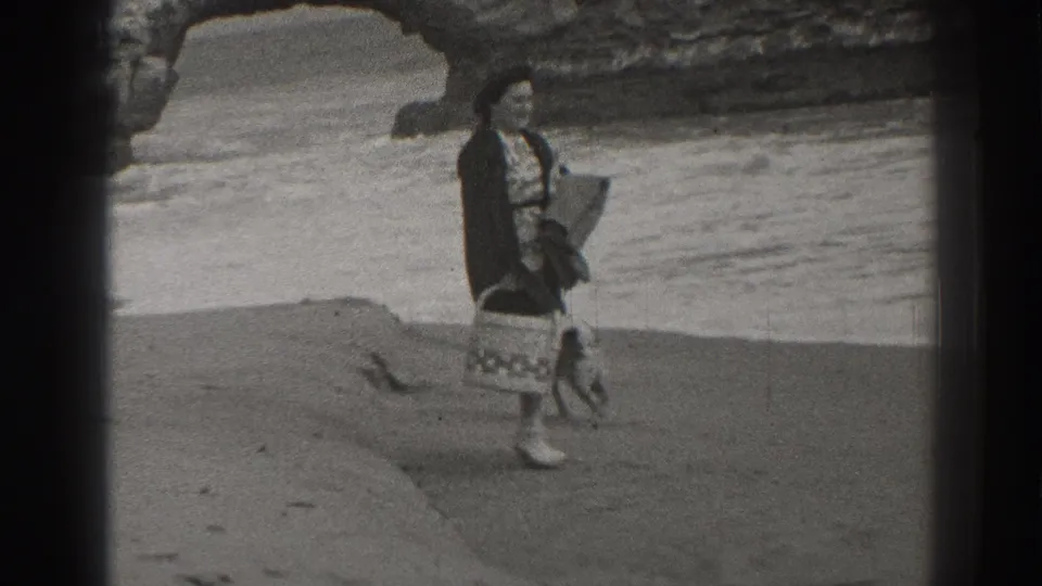 Woman in period beach attire carries a basket while a small dog stands nearby, with the Santa Cruz rock arch behind them.