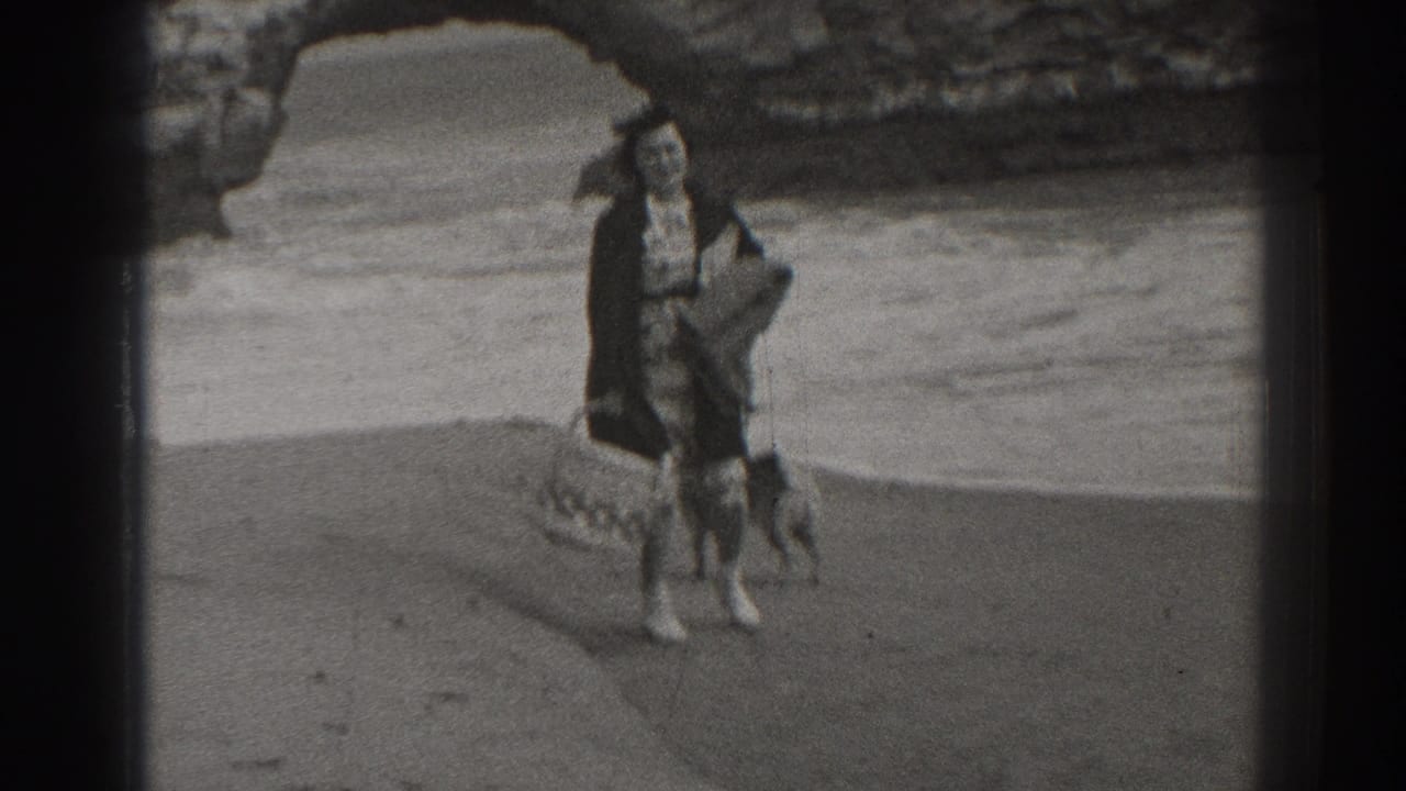 In 1937 Santa Cruz, a woman carrying a basket walks on the beach with two dogs near waves and a rocky arch.