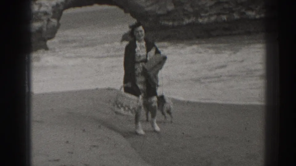 In 1937 Santa Cruz, a woman carrying a basket walks on the beach with two dogs near waves and a rocky arch.