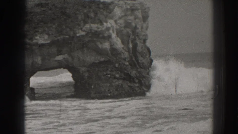 1937 Santa Cruz 8mm frame showing waves pushing through a rugged coastal sea arch in black and white.