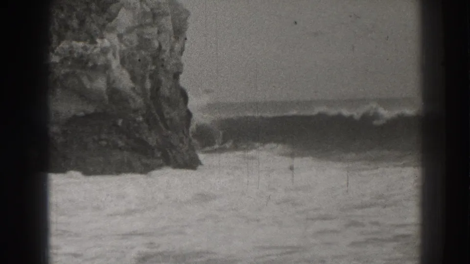 Black-and-white 8mm frame from Santa Cruz showing waves rolling beside a large coastal rock formation.