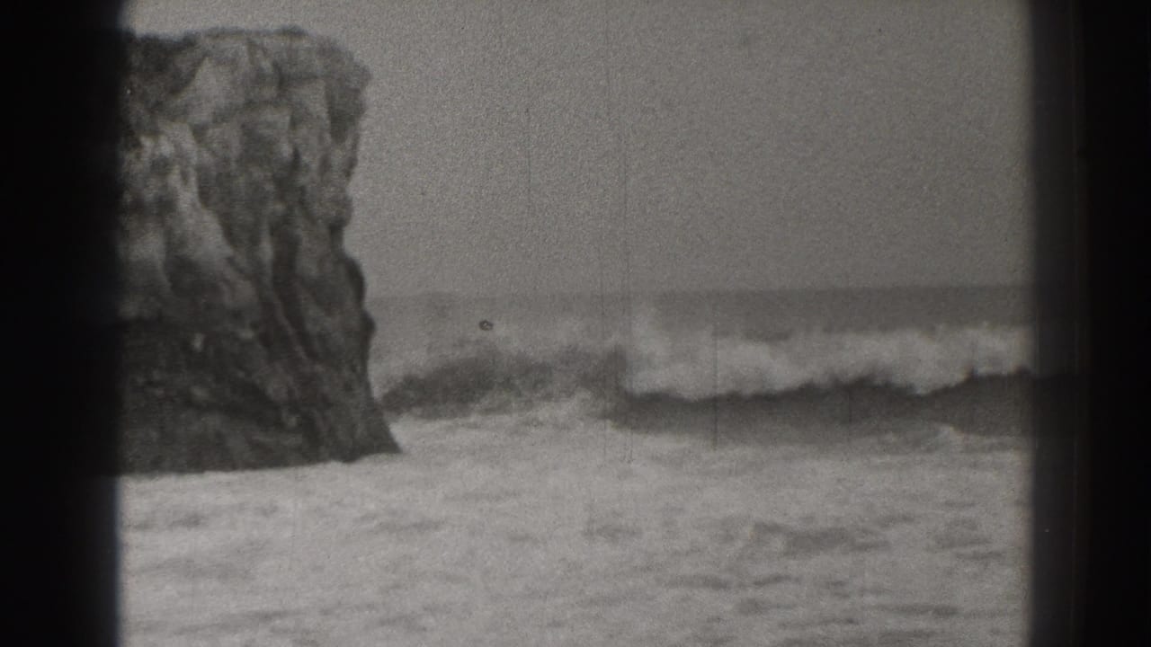 1937-era black-and-white 8mm view of a rocky California shoreline with surf breaking near a large sea stack.
