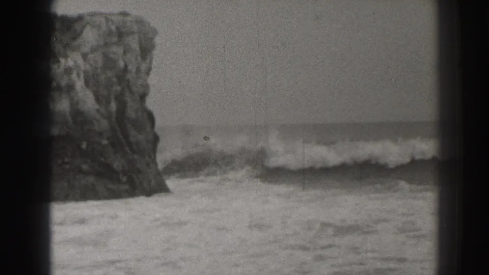 1937-era black-and-white 8mm view of a rocky California shoreline with surf breaking near a large sea stack.