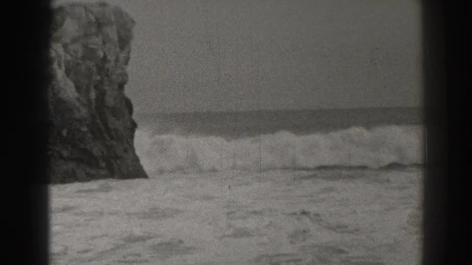Black-and-white 8mm frame showing waves moving past a large rocky formation on the California coast.