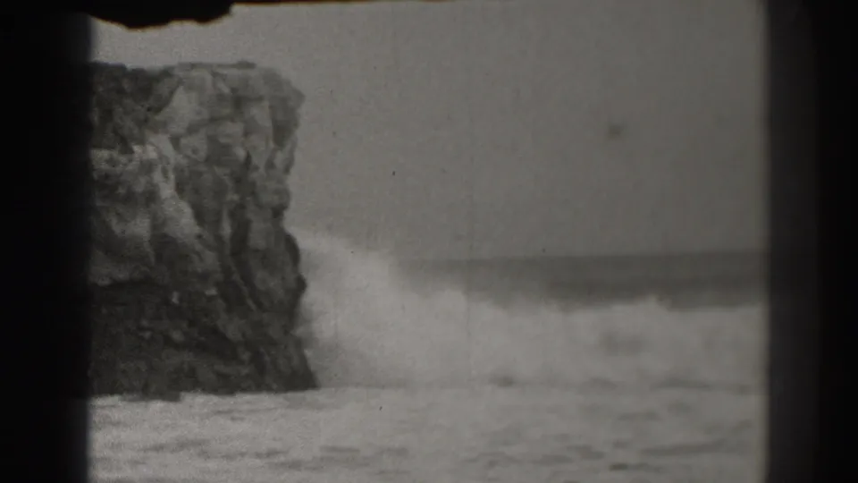 Soft black-and-white shoreline view with foamy water in front of a tall rock formation, consistent with 8mm home-movie footage.